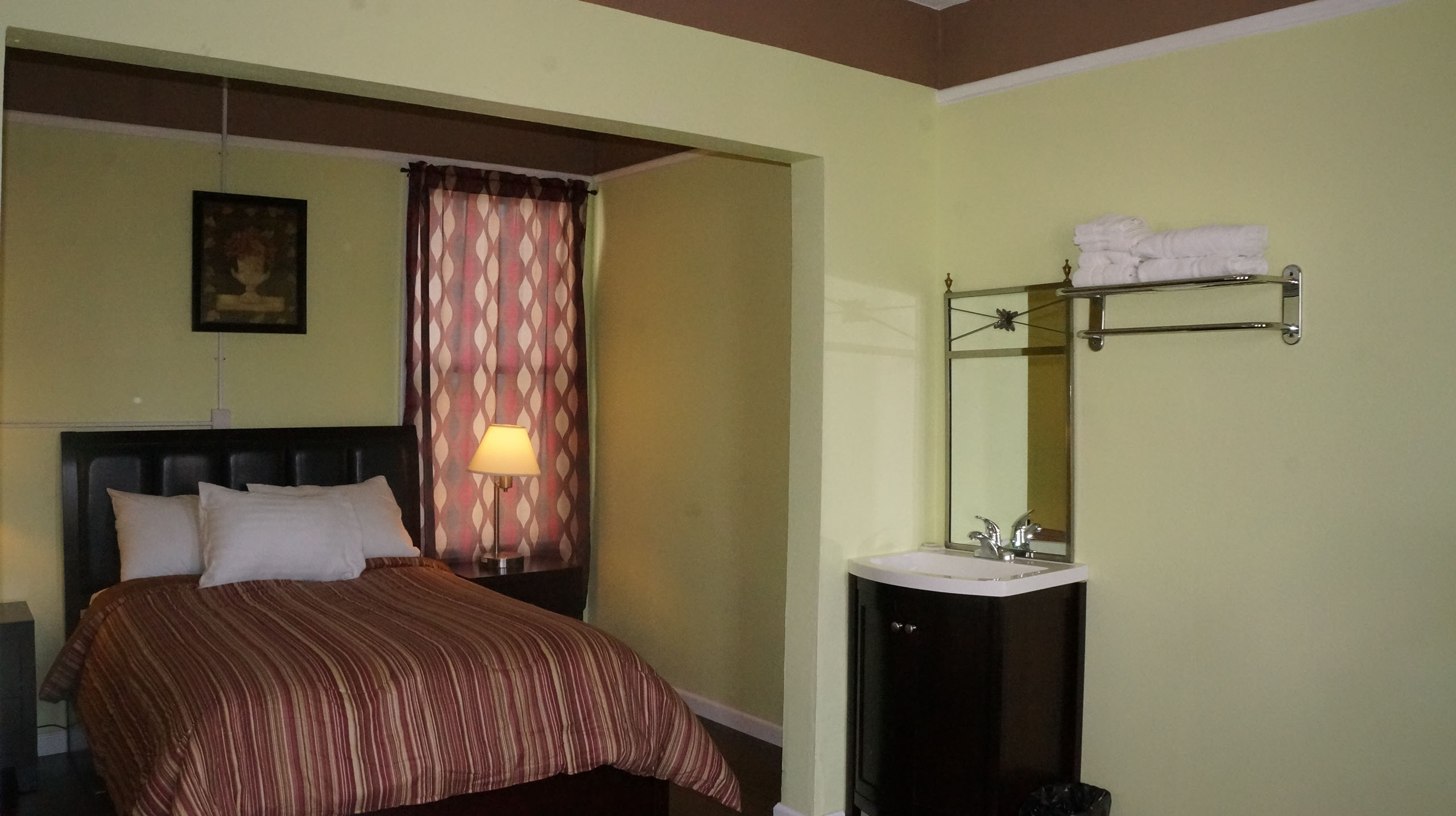 A hotel room with a bed, nightstand, lamp, window, sink, and mirror.