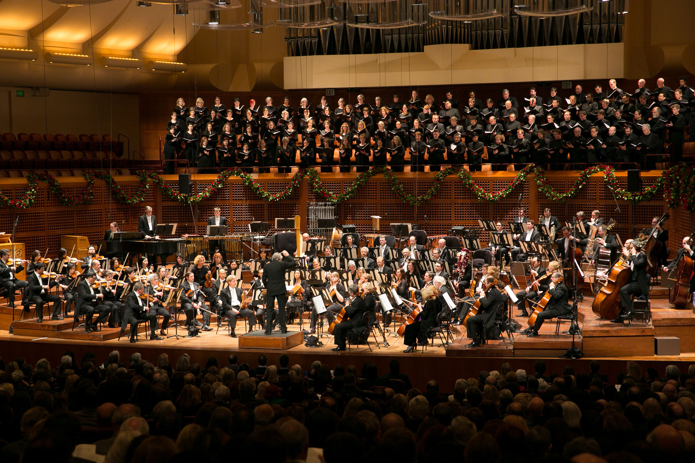 A large concert hall with an orchestra, choir, and conductor on stage, and a full audience seated in front.